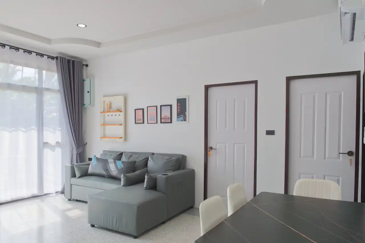 Shared living area at Baan Bahng Can Do Krabi homestay with L-shaped grey sofa, dining table, large garden-view window, air conditioning, and bedroom doors