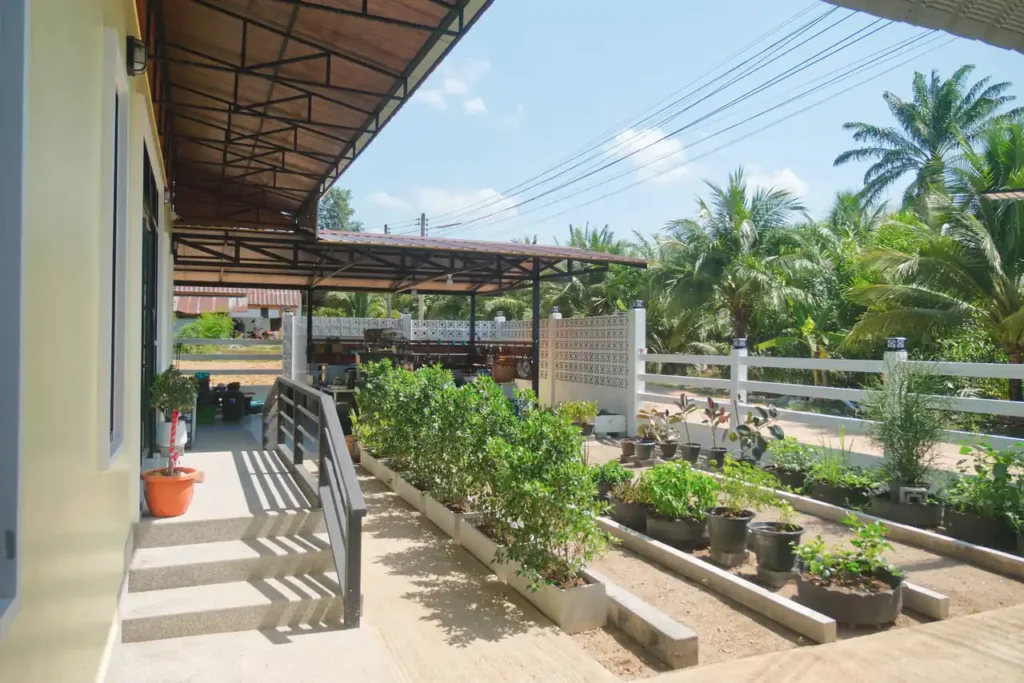 Front entrance path and herb garden at Baan Bahng Can Do Krabi homestay in Nong Thale, with raised plant beds, potted tropical plants, covered terrace, and palm tree surroundings