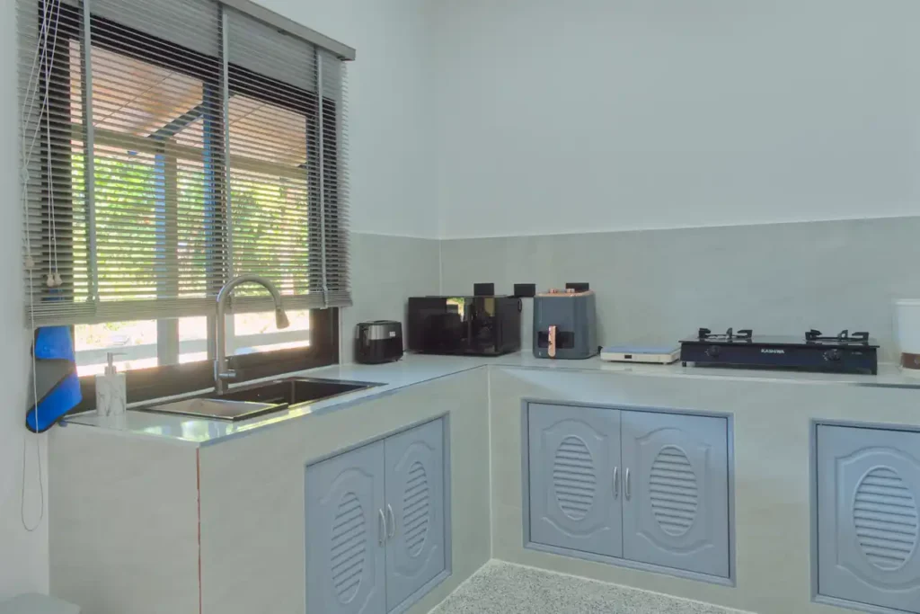 Indoor guest kitchen at Baan Bahng Can Do Krabi homestay with L-shaped countertop, sink, microwave, air fryer, toaster, and gas hob with garden window view