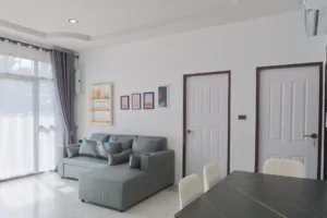 Bright shared living and dining area at Baan Bahng Can Do Krabi homestay with grey L-shaped sofa, large garden-view window with grey curtains, and dining table