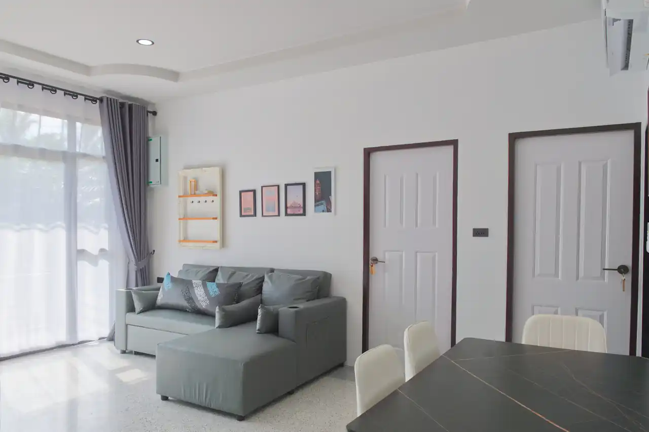 Bright shared living and dining area at Baan Bahng Can Do Krabi homestay with grey L-shaped sofa, large garden-view window with grey curtains, and dining table